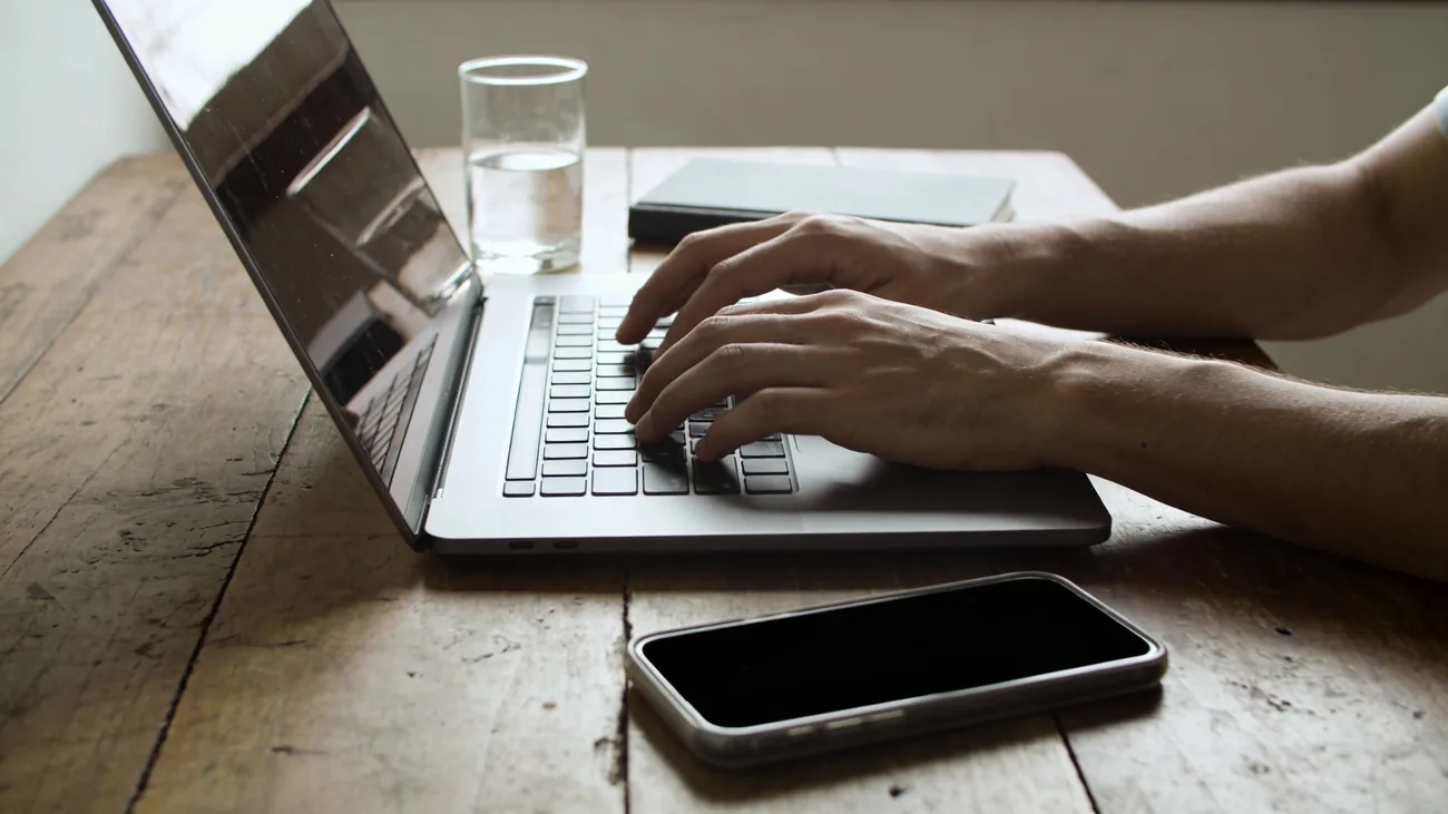 Mãos digitando em um notebook em uma escrivaninha de madeira em casa, smartphone com tela apagada ao lado, luz natural suave
