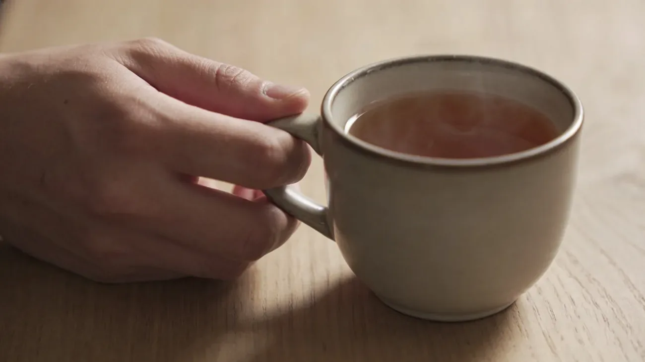A single hand holding a simple ceramic mug of warm tea on a tidy wooden surface, grounding atmosphere